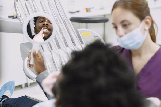 A man examines his teeth in a mirror during a dental check-up. A dentist is present.