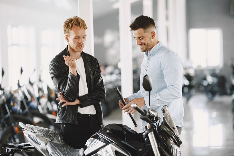 Two men engaged in conversation over a motorcycle in a showroom. Modern and vibrant setting.