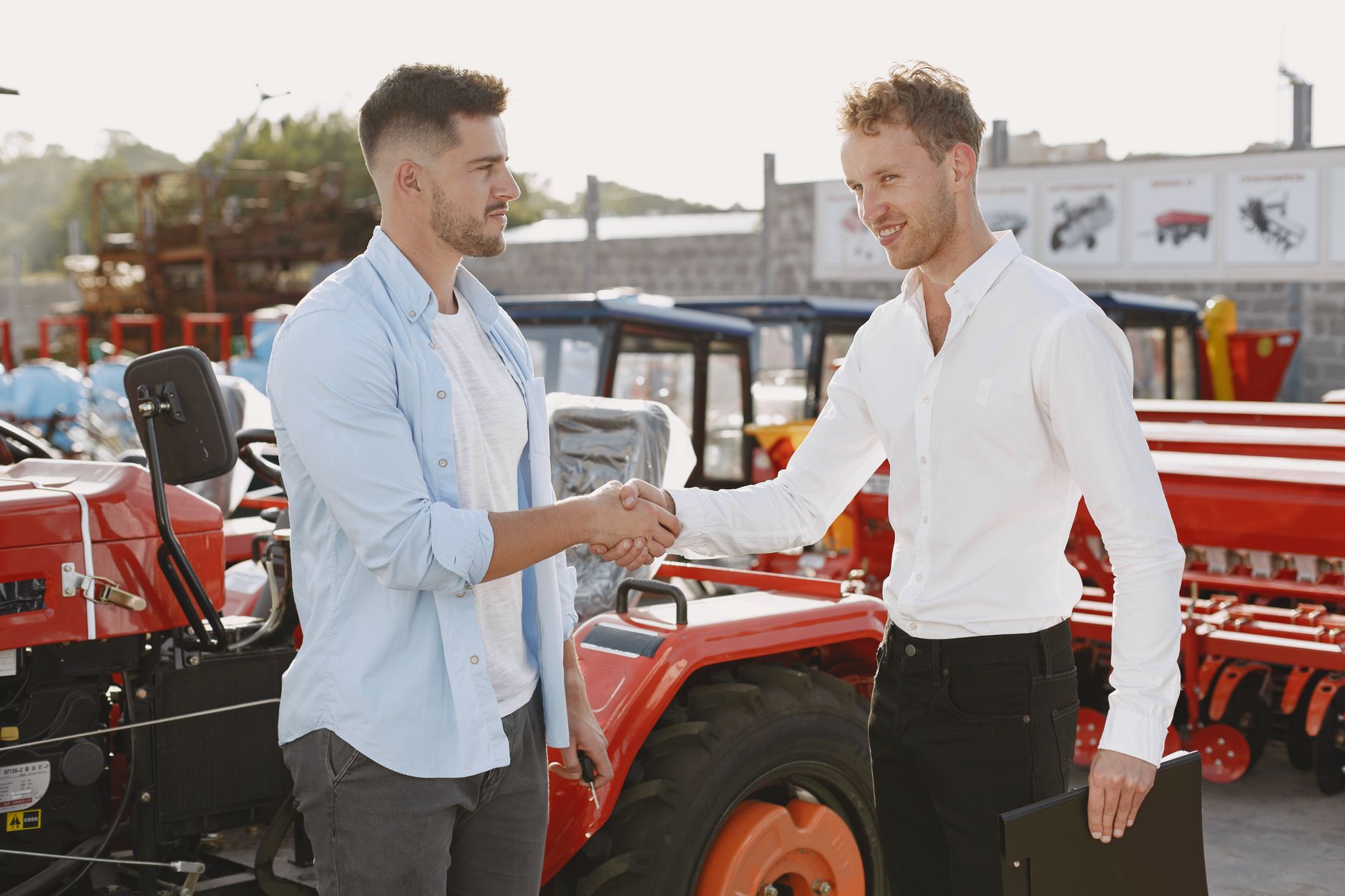 Two men shaking hands near a tractor, symbolizing a business deal or purchase agreement, outdoors.