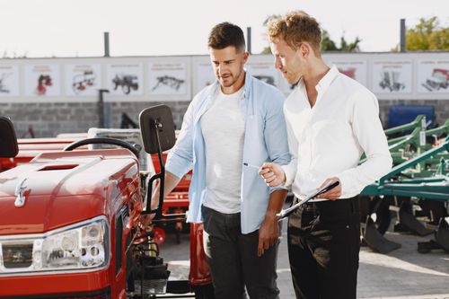 Free Two men having a discussion about a red tractor in an outdoor setting, possibly negotiating a deal. Stock Photo