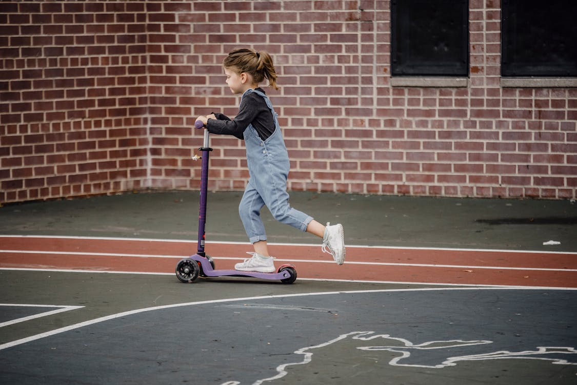Side view of excited preschool girl in casual outfit riding scooter on asphalt sports ground