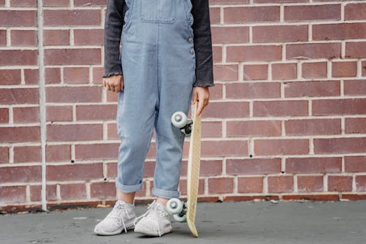 A child in denim overalls holds a skateboard against a brick wall, showcasing urban street style.