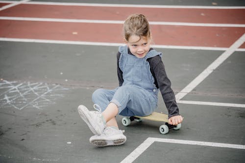 High angle of preschool  girl in casual outfit and sneakers sitting on skateboard while practicing skills in skate park
