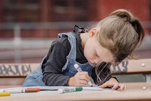 Free Adorable preschool girl in casual clothes sitting at table on schoolyard and holding marker while drawing illustration Stock Photo