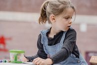 Crop cute preschool kid with ponytail in casual clothes sitting at table with colorful stationery for painting and looking away