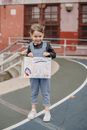 Full body of cheerful little girl in denim overall standing and showing drawing in hands and looking at camera