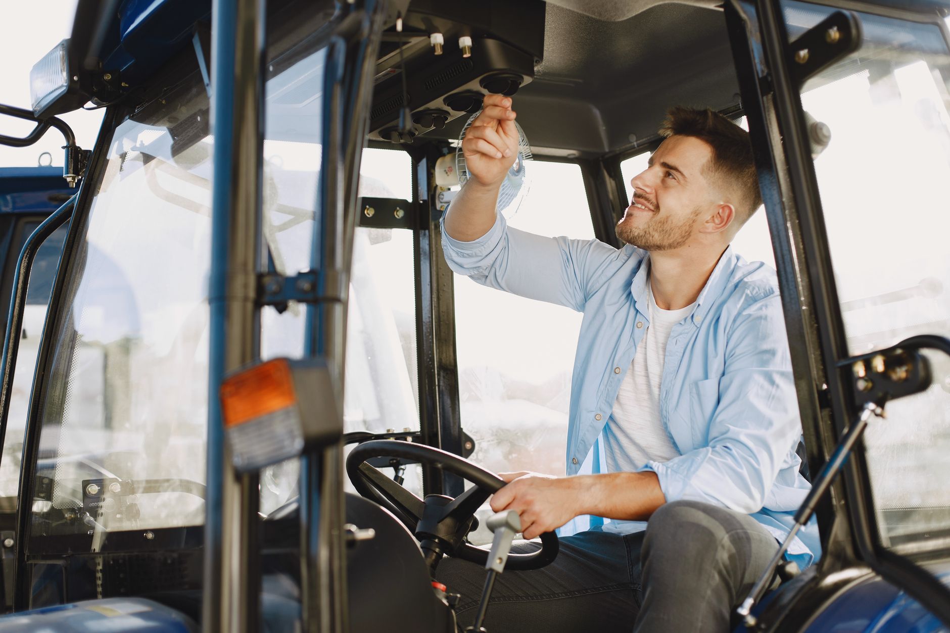 A man in a blue shirt checks controls while sitting in a tractor cabin outdoors.