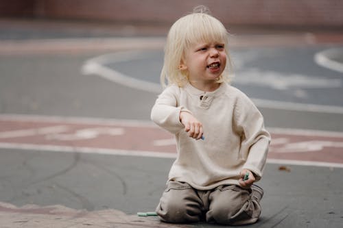 Free Full length dissatisfied little girl with chalks in casual wear sitting on asphalt ground and looking away Stock Photo