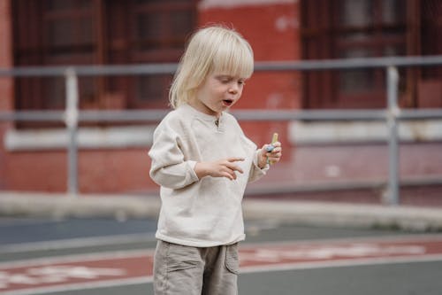 Cute little kid in casual wear standing on asphalt ground with chalks in hand and looking down ready to draw