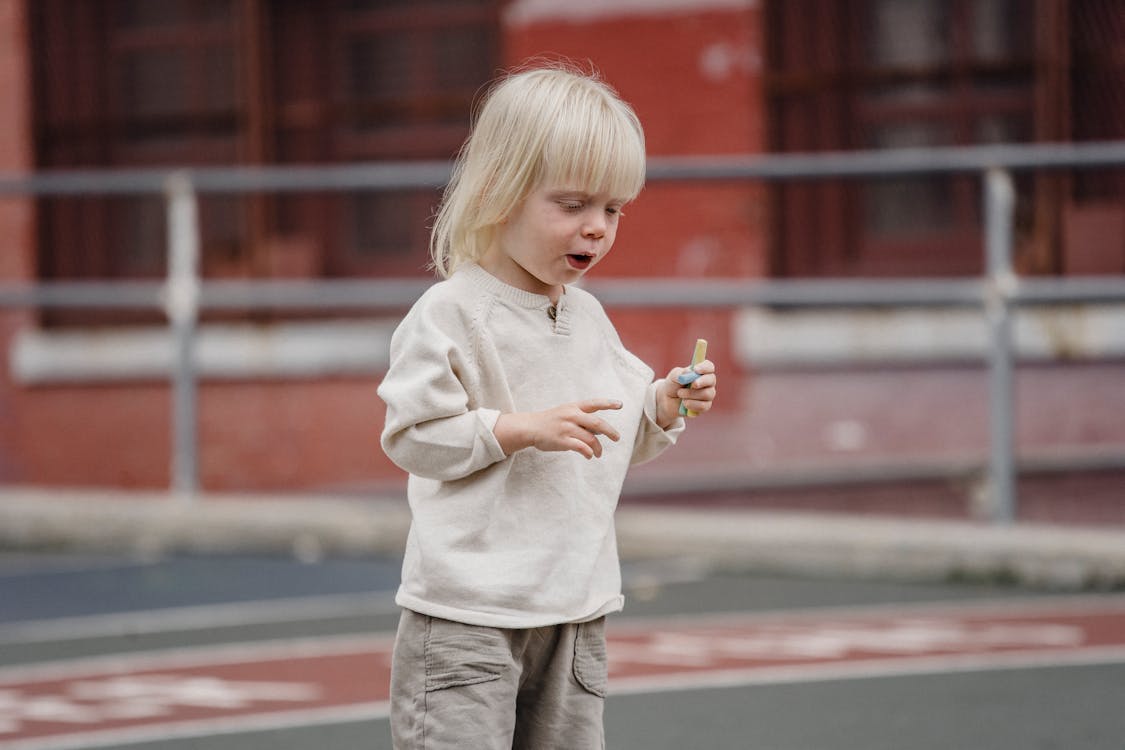 Free Cute little kid in casual wear standing on asphalt ground with chalks in hand and looking down ready to draw Stock Photo