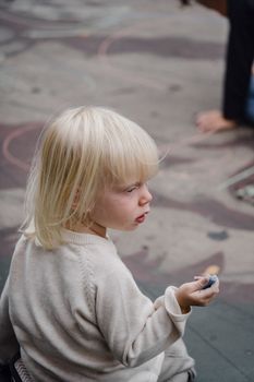从上面看，一个穿着休闲装的可爱小女孩坐在柏油地上，手里拿着彩色粉笔，目光移向别处