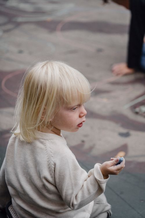 Free From above back view of cute little girl in casual wear sitting on asphalt ground with color chalk and looking away Stock Photo