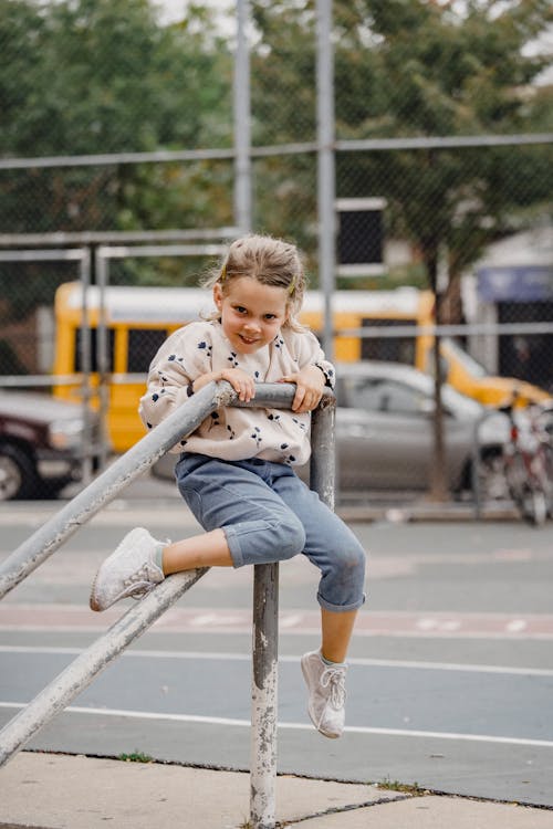 Full body delighted little girl in casual outfit sitting on metal railing and smiling while looking at camera