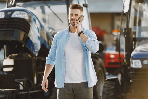 Free Confident young man wearing casual attire, speaking on phone outdoors near tractors. Stock Photo