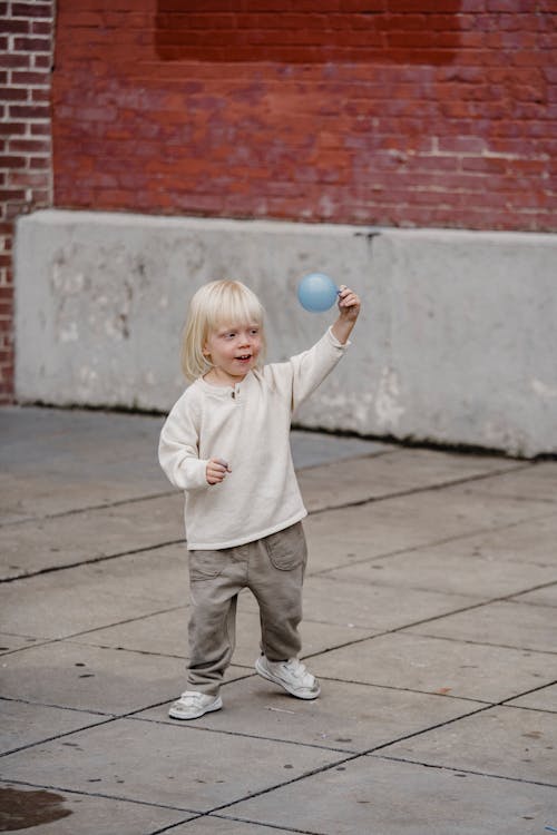 Full length happy cute boy wearing casual outfit raising hand with small balloon while standing on paved sidewalk