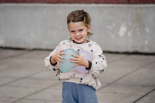 Sincere child in casual wear with ornament looking at camera with assorted balloons on city street