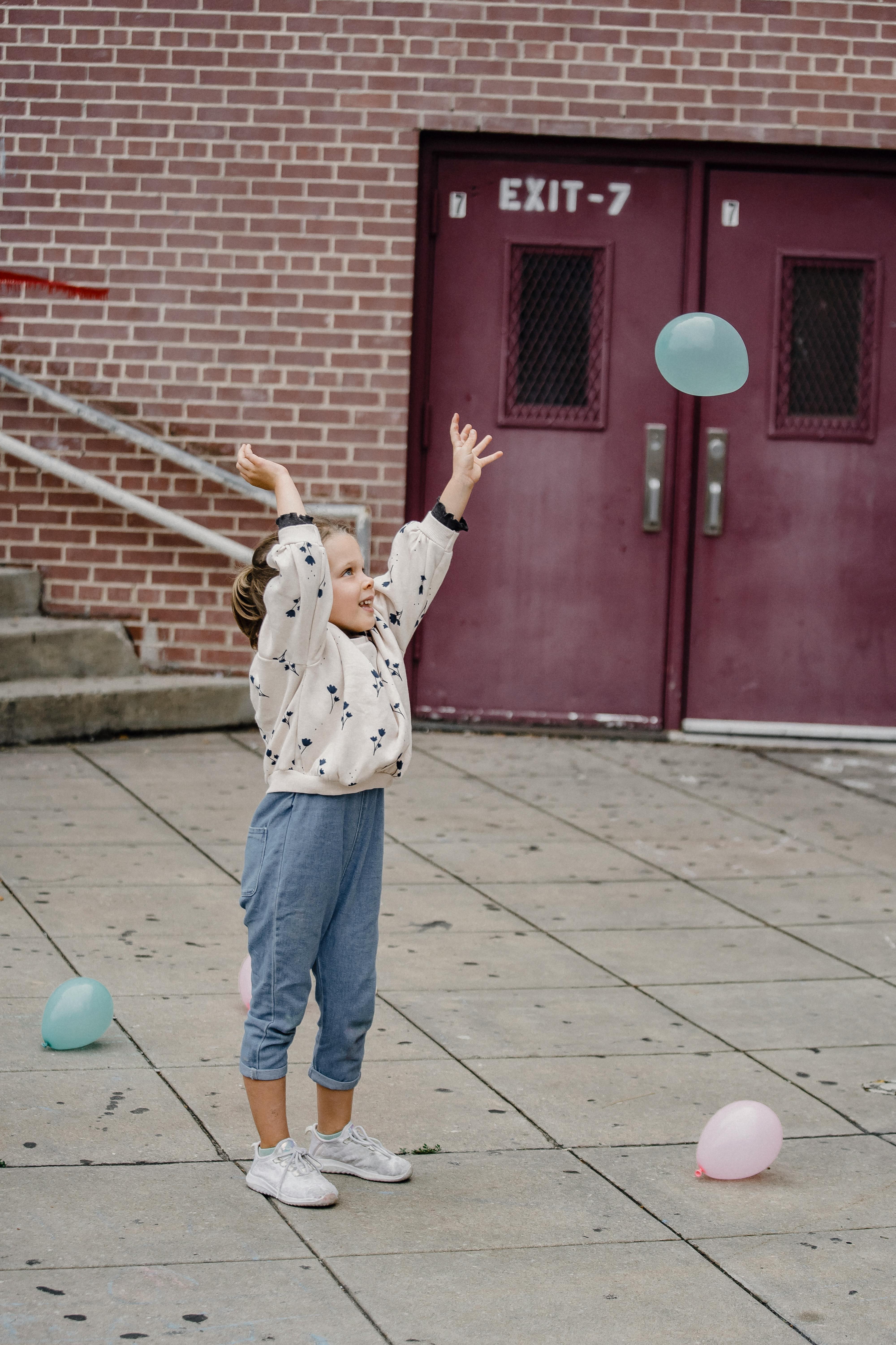 Happy child in casual wear joyfully throwing balloons outside a brick building.