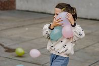 Kid in ornamental apparel with barrette on hair strolling on street with different balloons while looking forward