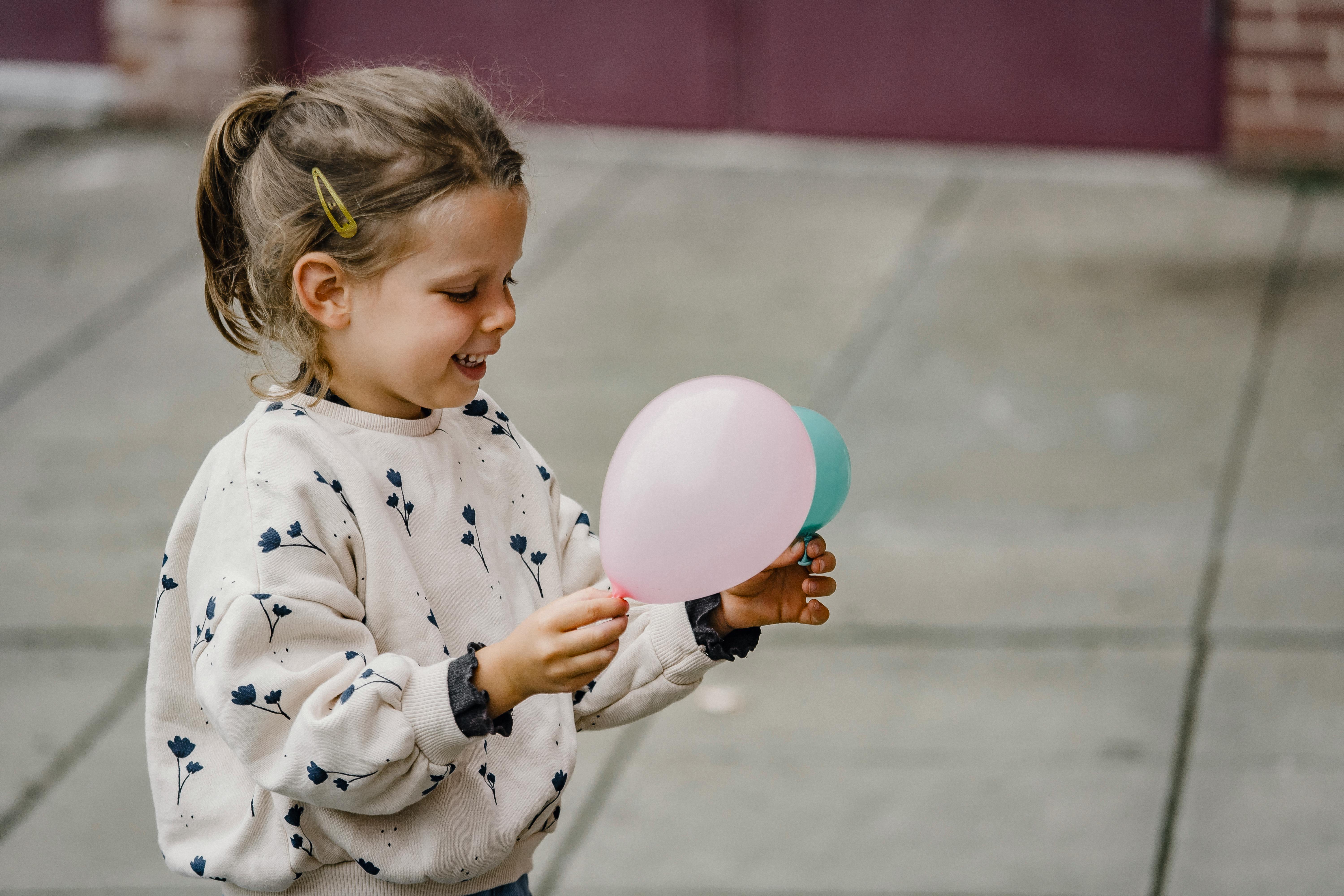 Side view of glad child in casual clothes with ornament playing with balloons on urban street in daylight