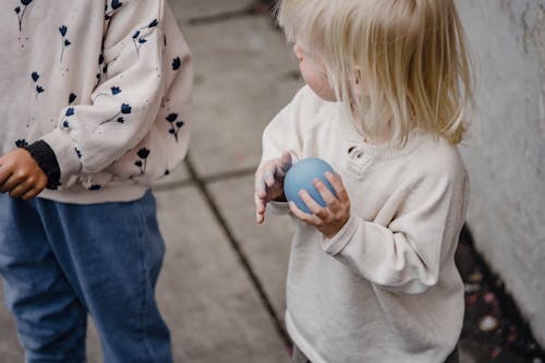 High angle of crop blond child with small balloon spending time with anonymous friend in informal clothes on pavement in daylight