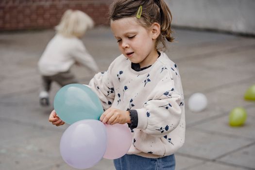 Crop charming child in ornamental apparel standing with assorted balloons on street with anonymous best friend playing in daytime