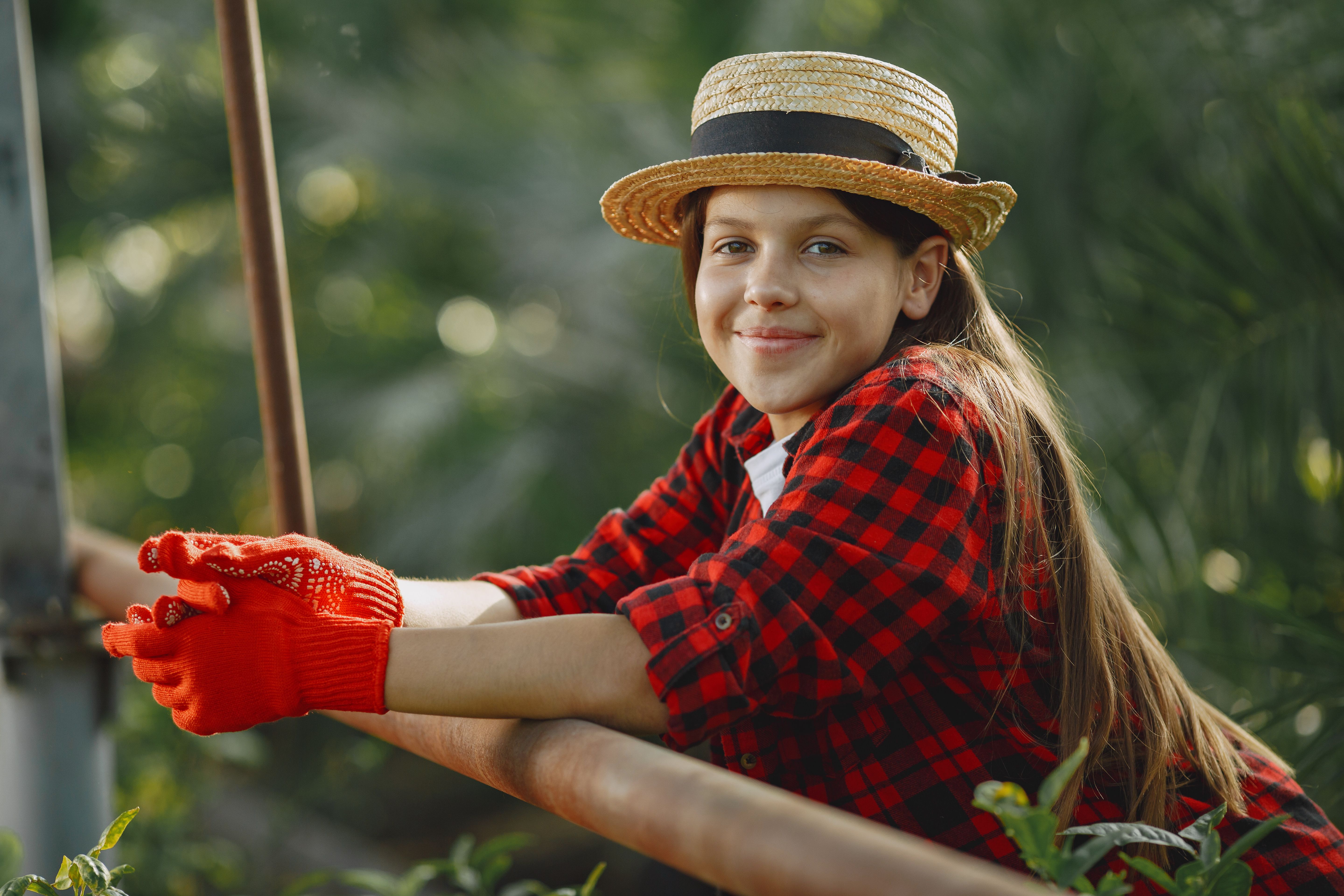 A cheerful young girl wearing a straw hat and plaid shirt smiles while enjoying an outdoor environment.