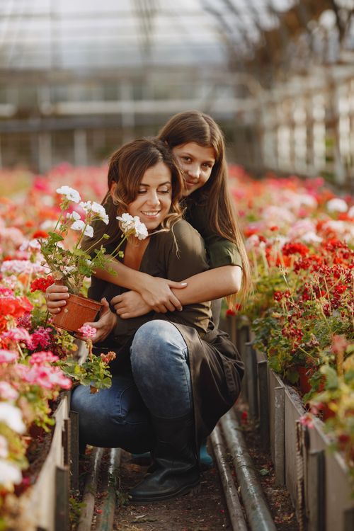 Free A mother and daughter share a joyful hug surrounded by vibrant flowers in a greenhouse. Stock Photo