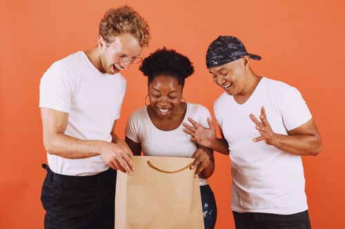 Free Three diverse adults joyfully open a paper shopping bag against an orange backdrop, showcasing multicultural friendship. Stock Photo