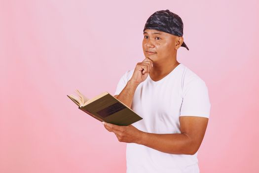 A thoughtful man in a cap and white t-shirt reads a book against a pink background.