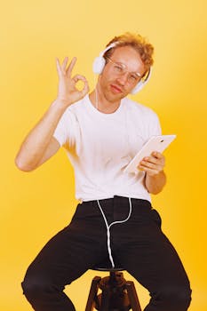 Adult man wearing headphones and using a tablet, giving an OK hand gesture against a yellow background.