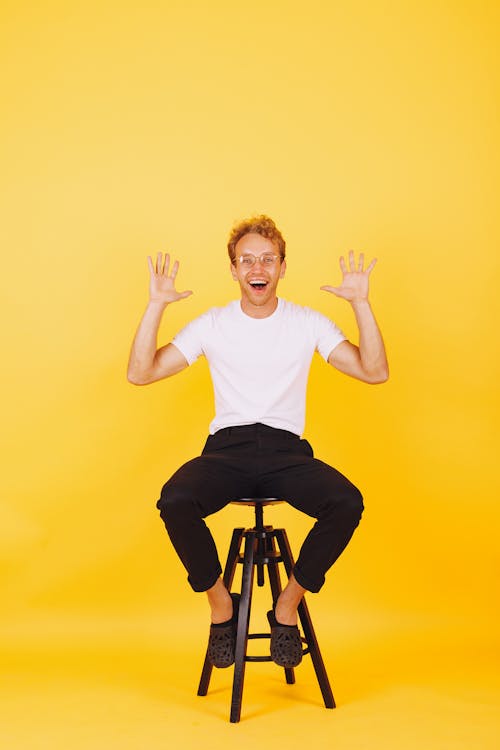 Man in White Crew Neck T-shirt and Black Pants Sitting on a Stool With Hands Up