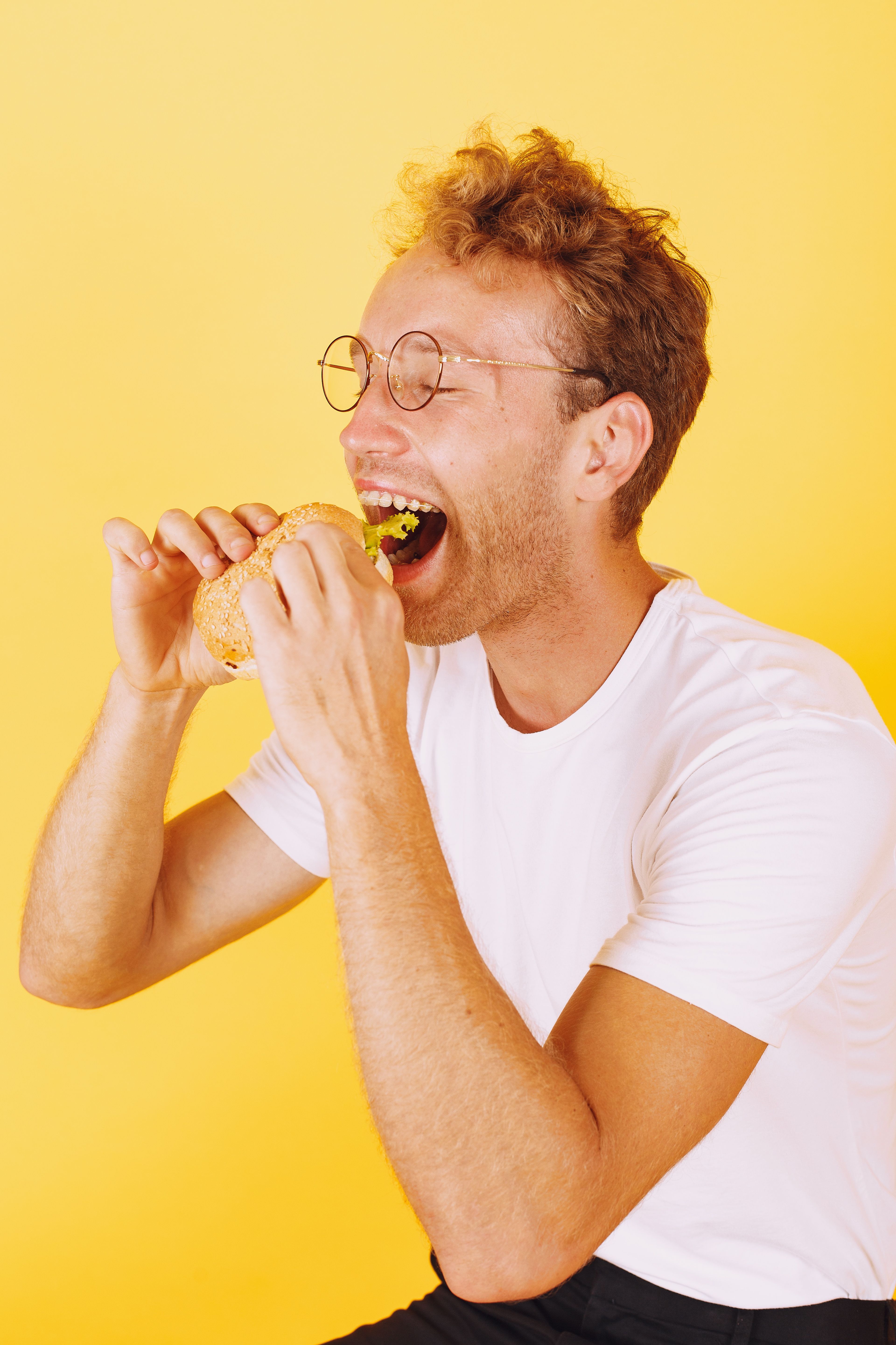 Caucasian man in glasses eating a delicious burger against a vibrant yellow background.