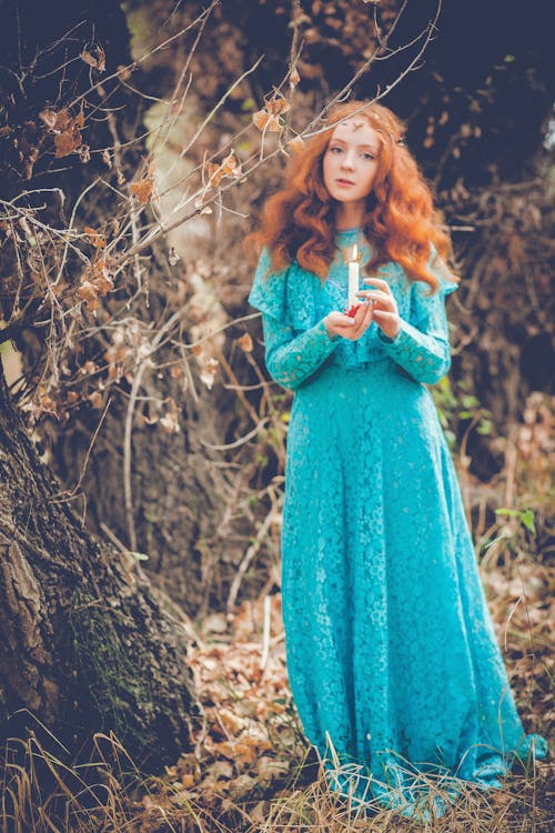 Young contemplative lady with red hair in bright elegant dress standing with burning candle near trees while looking at camera