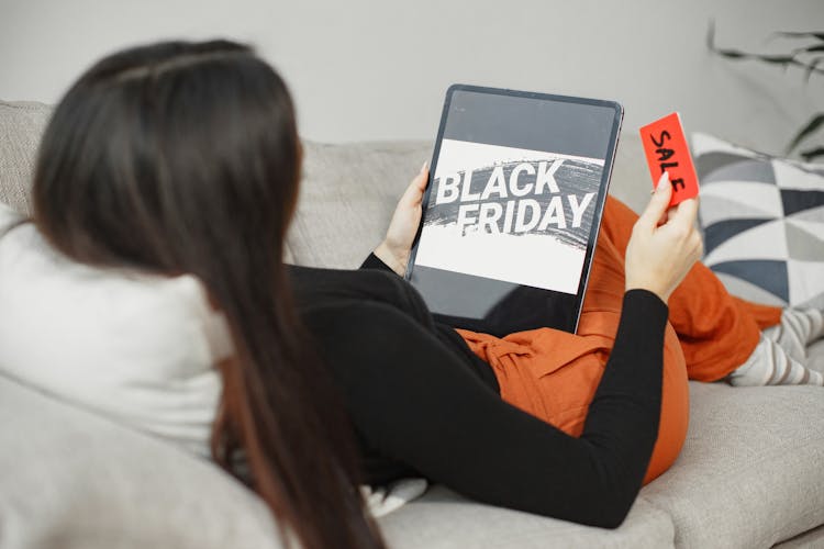 Woman Lying On A Couch And Holding A Tablet Displaying A Black Friday Sale 