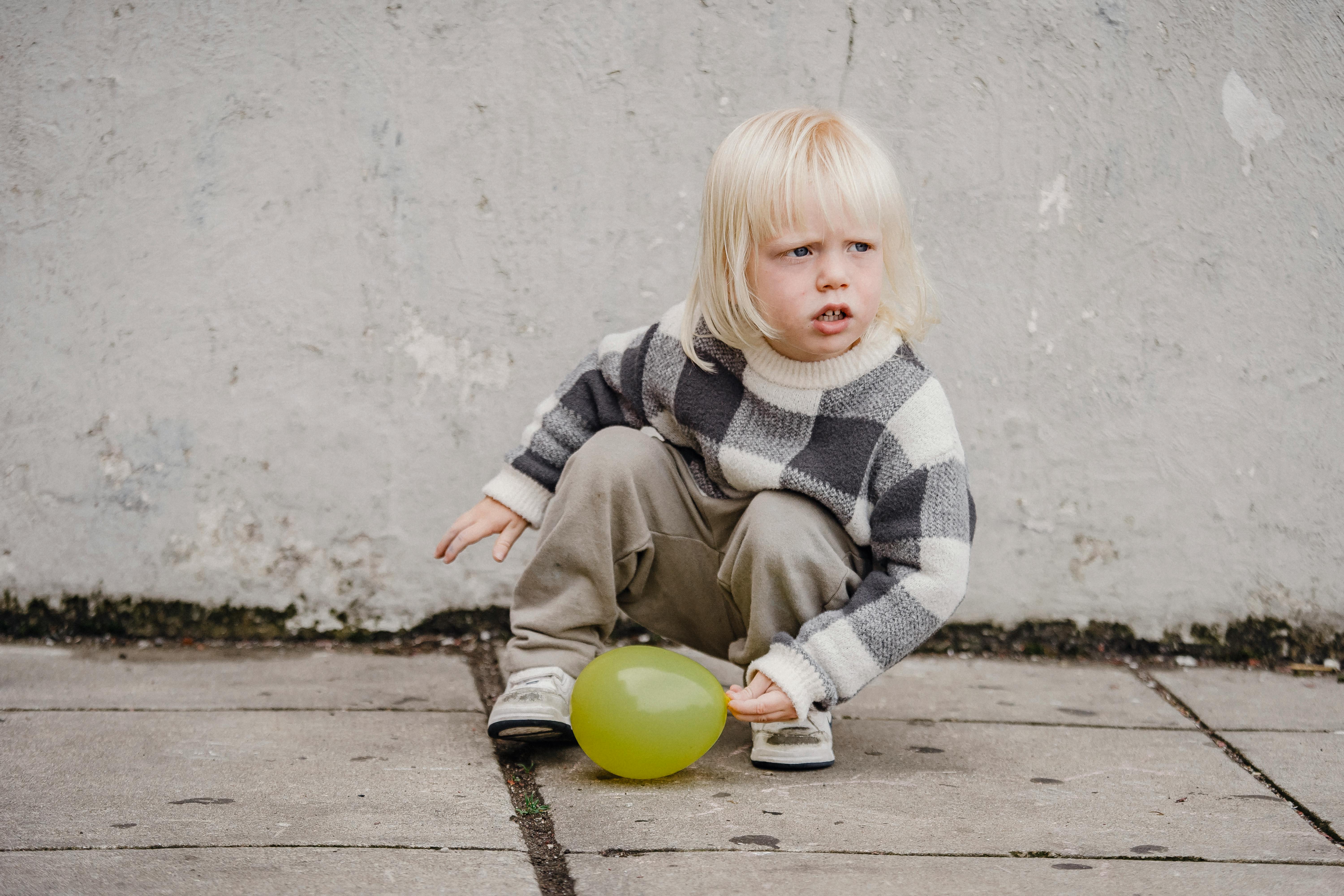 Adorable attentive child in stylish outfit squatting with balloon on pavement and looking away