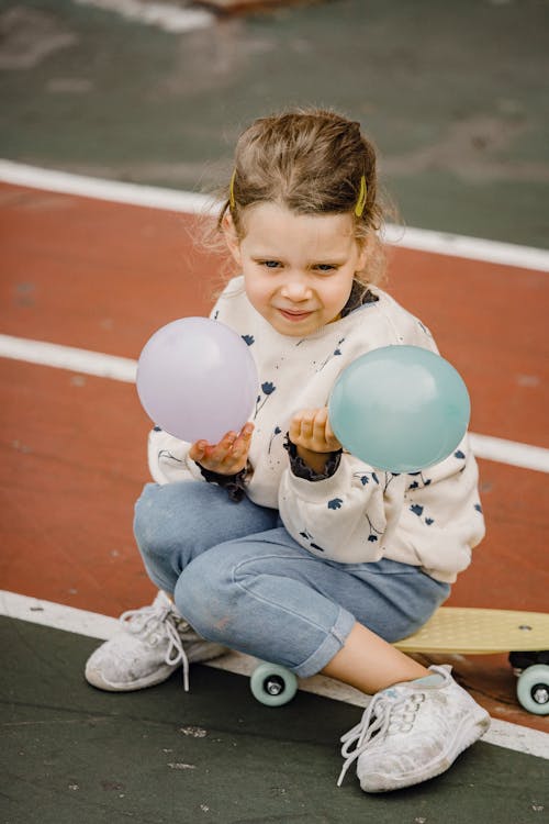 Free From above of sincere child in informal outfit with balloons sitting on skateboard and looking forward on sports ground Stock Photo