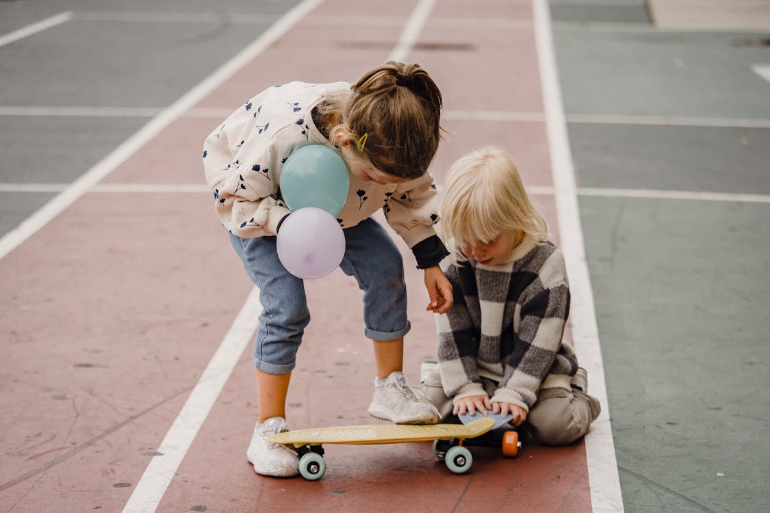 Free Unrecognizable kids in casual outfit interacting while standing on pavement with balloons and skateboard in city Stock Photo