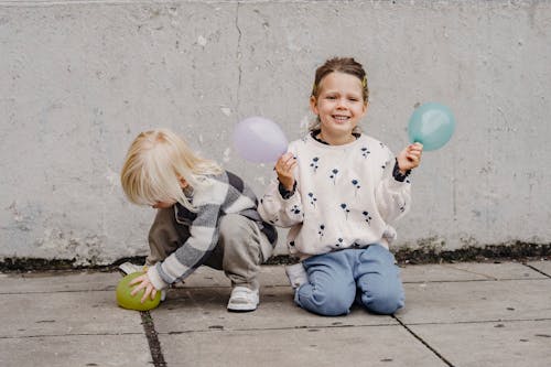 Cheerful child in informal wear sitting on pavement with balloons near anonymous little kid against cement wall