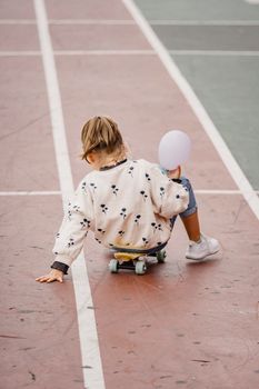 白天，一位身穿休闲装、手拿气球的匿名女孩，背对镜头坐在公路长板上的全身背影