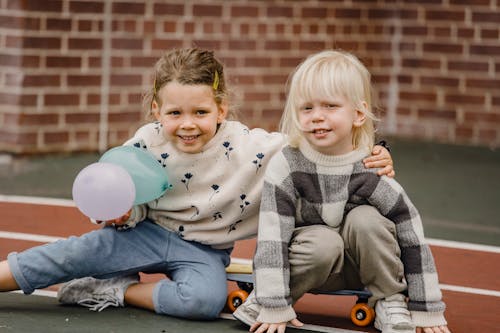 Happy children in casual outfit with balloons sitting on skateboard on road near wall in daytime