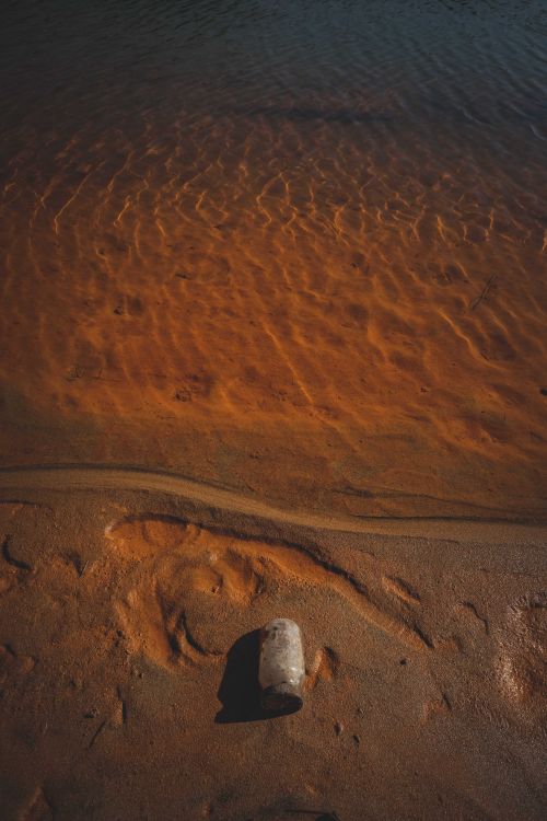 Free From above of clear sea washing sandy shore with traces and thrown plastic bottle in sunlight Stock Photo