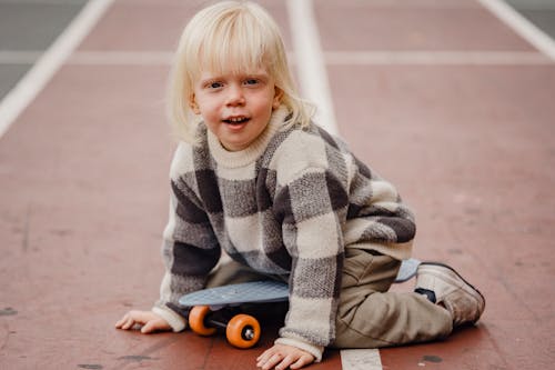 Full body of playful little kid looking at camera while sitting on sports ground with penny board against blurred background