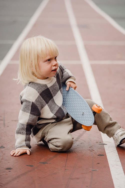 Side view of little kid wearing checkered sweater sitting on ground of playground with penny board in hand against blurred background