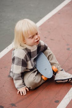A smiling blond child sitting on a sports track with a skateboard, enjoying a playful moment outdoors.