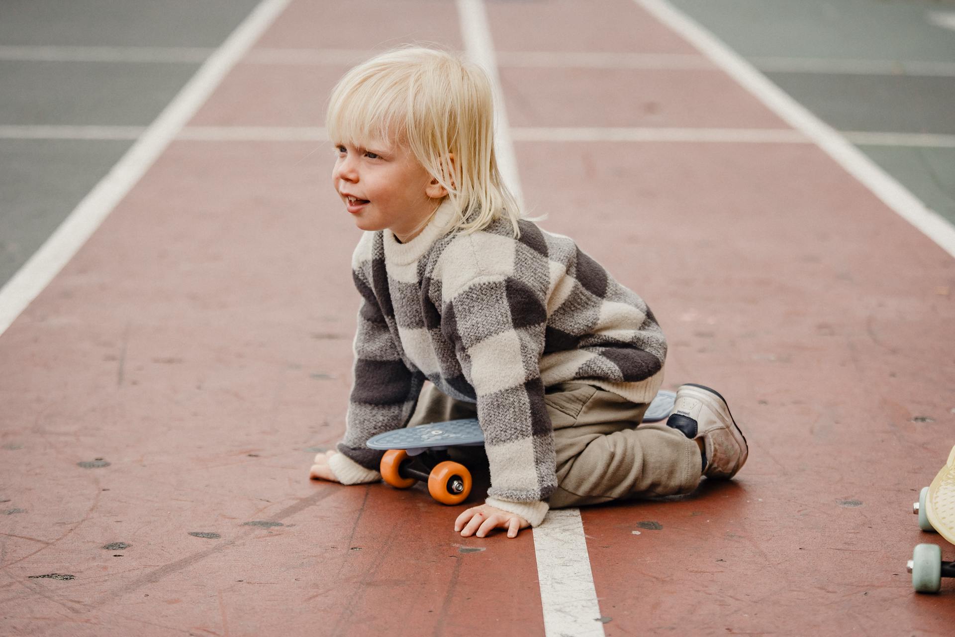 Full body side view of playful little kid with penny board sitting on red track of sports ground on blurred background