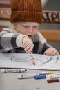 Crop concentrated little kid wearing hat sitting on wooden bench at table while drawing with red felt pen on white paper sheet on street against blurred background