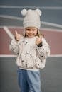 Little girl in jeans white jumper and hat showing thumbs up standing in playground and looking at camera