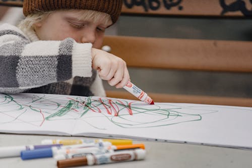 Little blond kid drawing in sketchpad using colorful markers sitting at table on playground