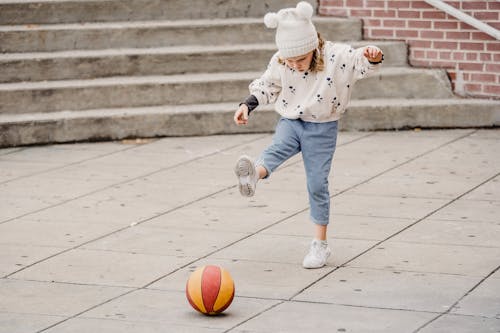 Full body of focused little girl in jeans white jumper and hat kicking colorful ball on street