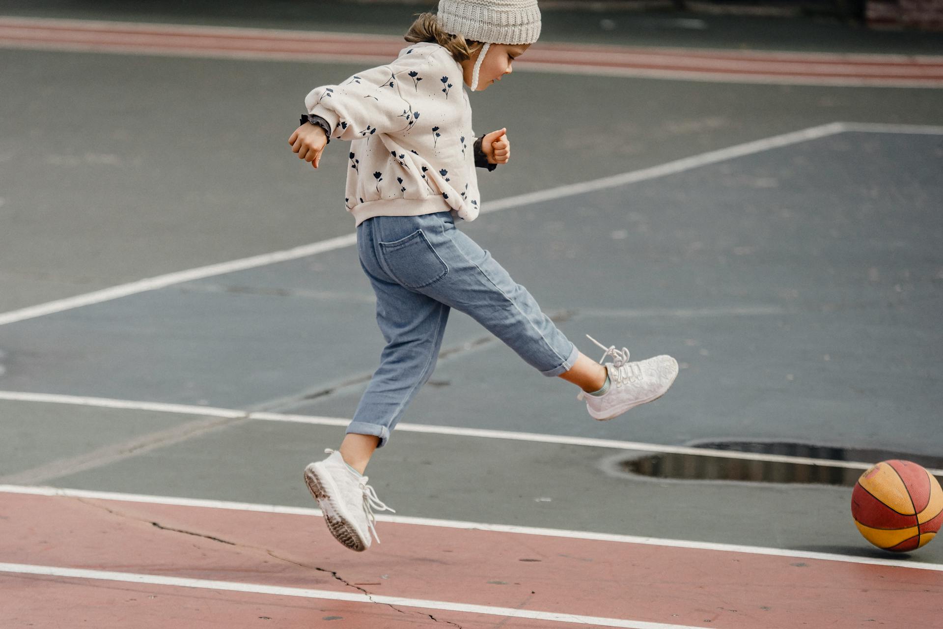 Side view of jumping little girl in jeans and jumper kicking colorful ball playing on sports yard